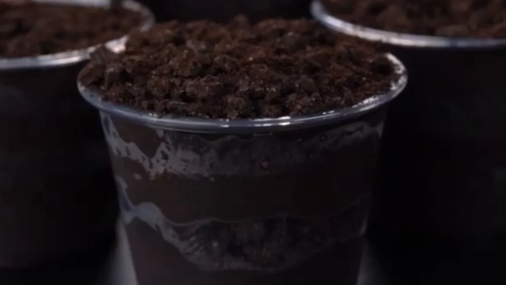 A close-up view of three transparent cups layered with glossy, dark brown chocolate protein pudding and crumbly, near-black crushed Oreo toppings. The distinct layers alternate between the smooth sheen of the pudding and the matte, soil-like texture of the Oreos.